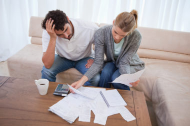 An image of a frustrated couple looking over many bills spread across their coffee table.